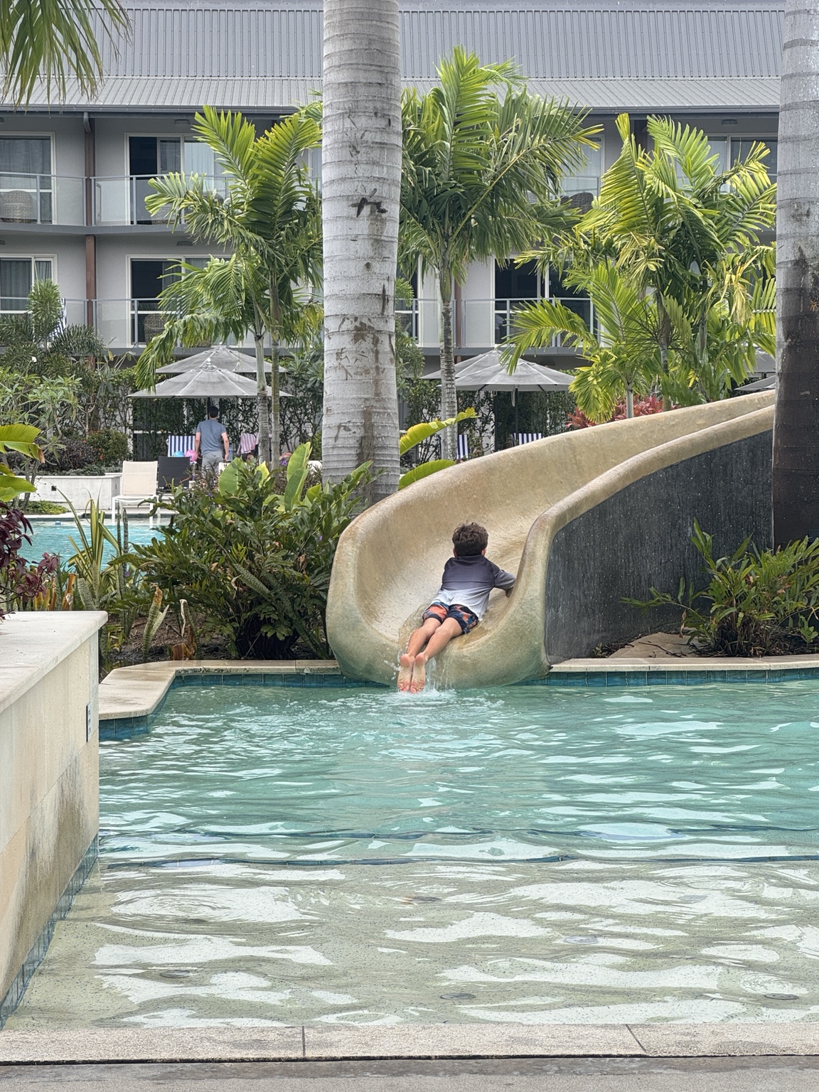 Child seen from behind using the small slide into Crowne Plaza Nadi’s children’s pool, with palms and accommodation blocks behind